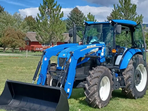 A blue New Holland tractor with a front loader attachment parked on a grassy field, with trees, and a blue sky at Heber Valley Cheese